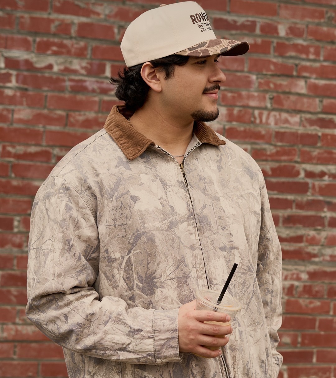 Model in a camo jacket and cream cap against a brick wall