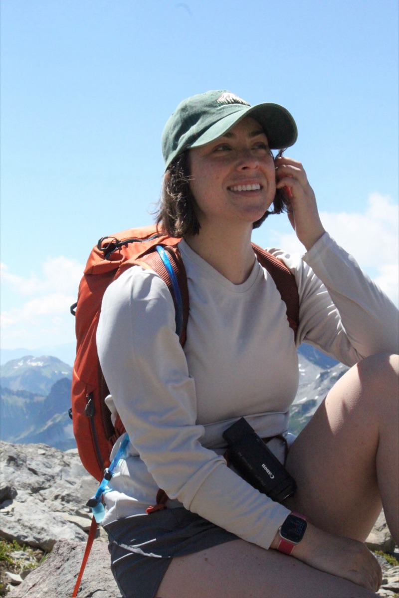 Makenna smiling on a rocky summit at Mount Rainier, wearing a green cap and an orange backpack