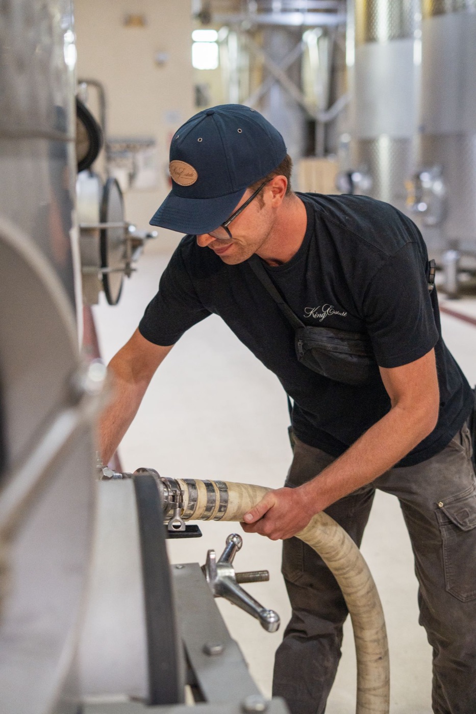 Winemaker in a King Estate cap connecting a hose to a stainless steel tank